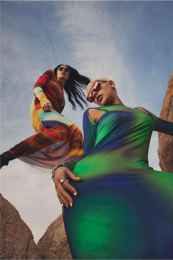 Two women pose in colorful long-sleeve dresses and sunglasses in a desert landscape under a partly cloudy sky, fashion photoshoot.