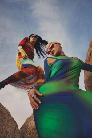 Two women pose in colorful long-sleeve dresses and sunglasses in a desert landscape under a partly cloudy sky, fashion photoshoot.