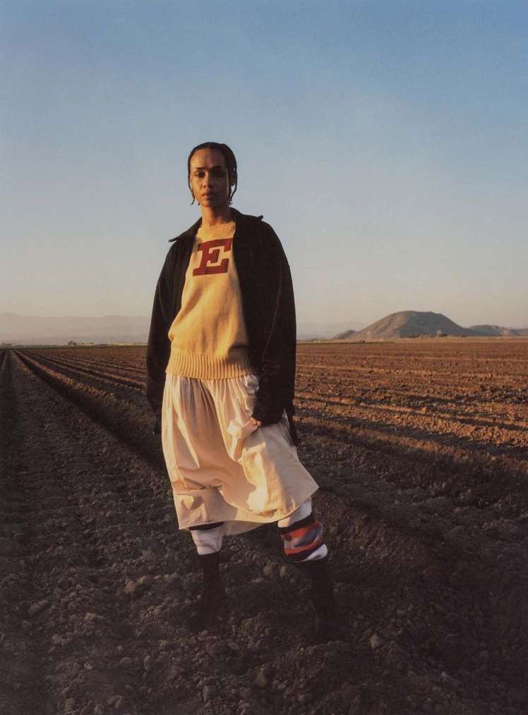 A woman stands in a field wearing a sweater, skirt, jacket, sneakers, and socks. The field stretches toward a distant mountain range.