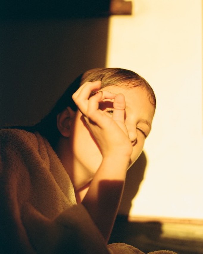 Portrait of a young person with a towel around their shoulders, partially obscuring their face with a hand in warm, directional light.