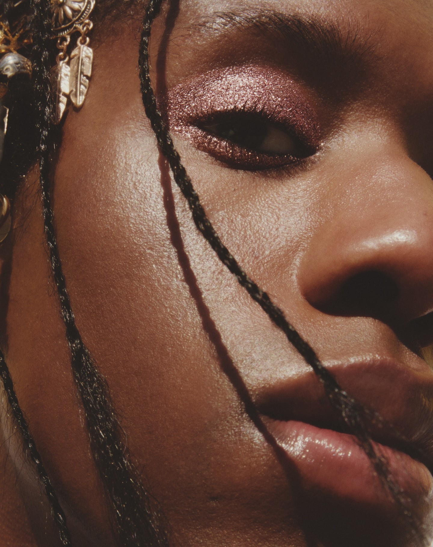 Close-up of a dark-skinned adult female face, featuring metallic rose eye makeup and ornate gold jewelry in braided hair.