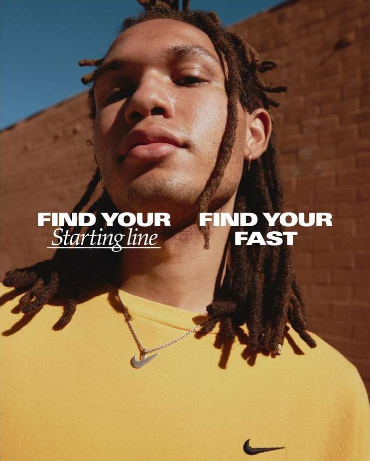 Close-up of an adult male with dreadlocks wearing a yellow shirt and necklace, posing in front of a brick wall under a blue sky.