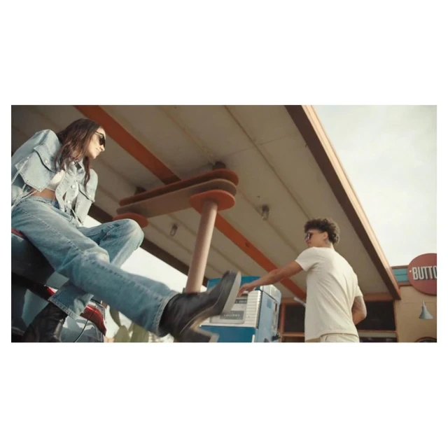 Two young adults in casual clothing at a gas station: a woman sits on a car and a man uses a gas pump, under the station's canopy.