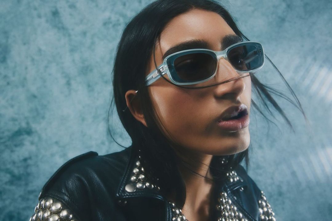 Close-up of an adult woman with sunglasses wearing a studded black leather jacket against a textured light blue backdrop.