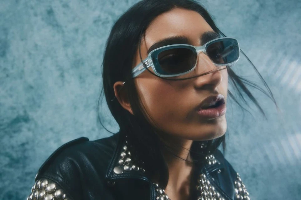Close-up of an adult woman with sunglasses wearing a studded black leather jacket against a textured light blue backdrop.