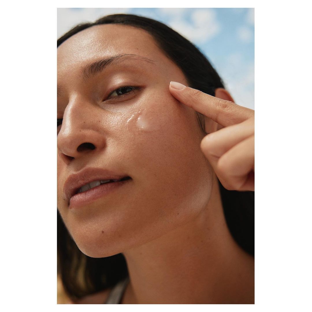 Close-up of an adult woman applying a drop of skincare product to her cheek against a sky background for beauty advertising.