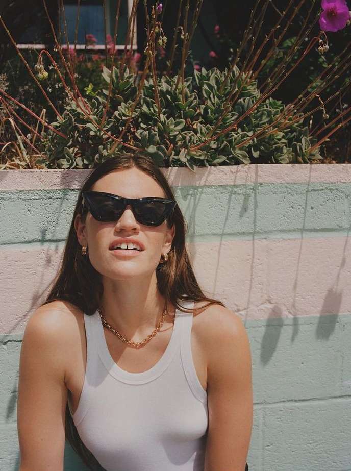 A woman poses outdoors wearing sunglasses and a white top with a green and pink wall and plants in the background on a sunny day.