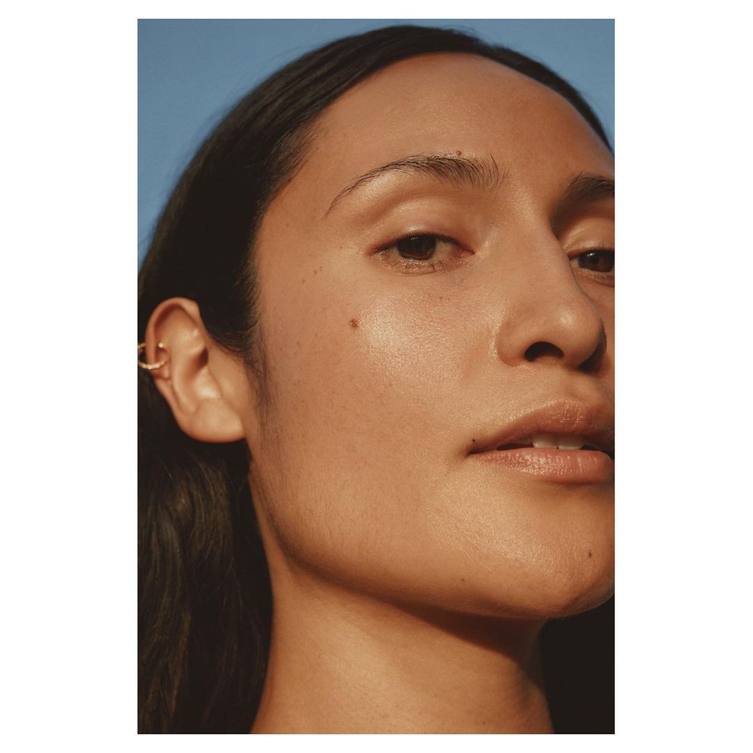 Close-up portrait of a woman profile showing her face, skin texture, and a gold ear cuff against a blue background by Ward and Kweskin.