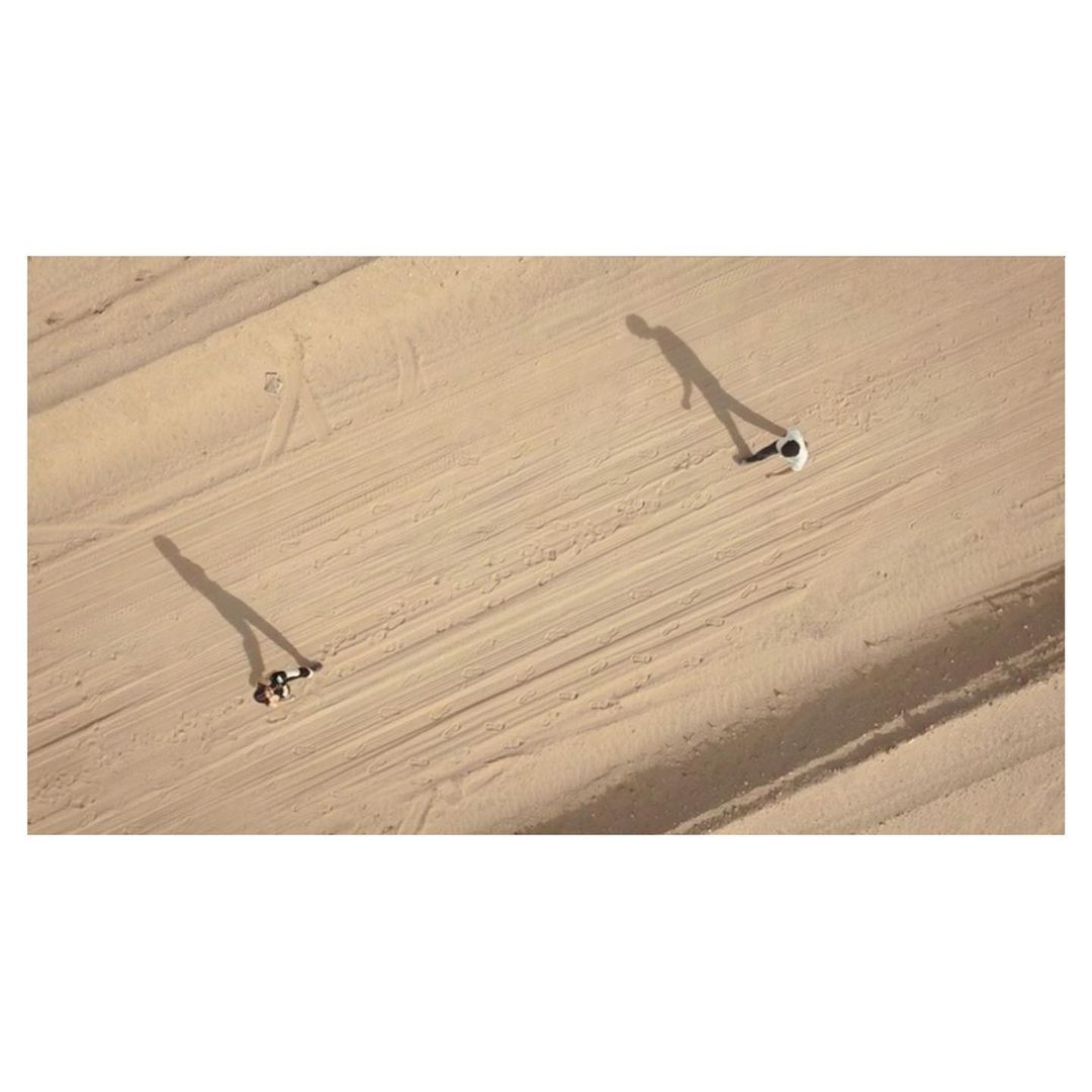 Aerial view of two people in a desert-like environment; one is walking while the other is on a sandboard, creating long shadows.