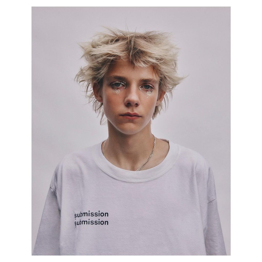 Portrait of a young model with messy blonde hair and glitter makeup wearing a white t-shirt against a plain grey background