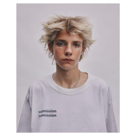 Portrait of a young model with messy blonde hair and glitter makeup wearing a white t-shirt against a plain grey background