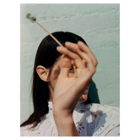Close-up portrait of a woman holding a thin stem with a small blossom while partially obscuring her face with her hand.
