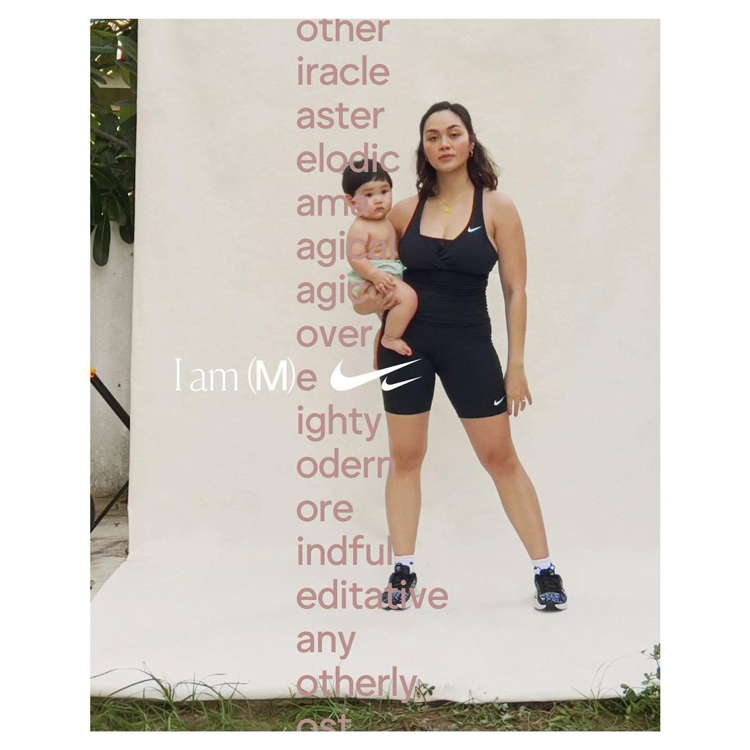 Mother holds her baby against a branded backdrop in a fashion-style shot. She's wearing black athletic wear and tennis shoes.
