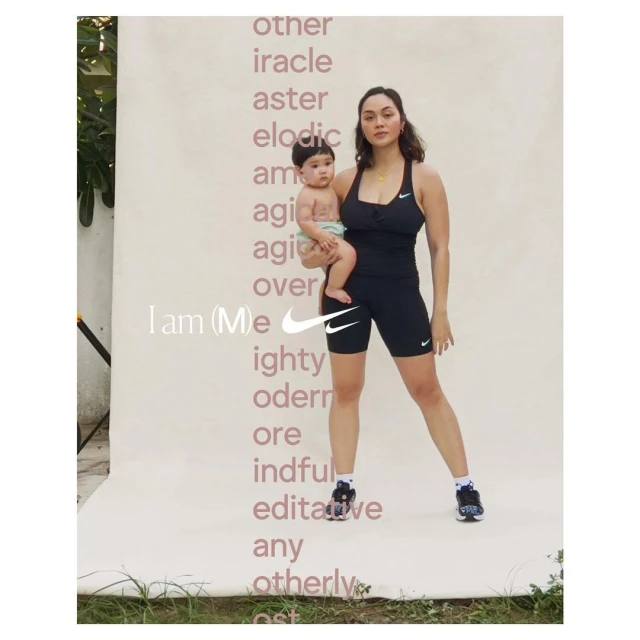 Mother holds her baby against a branded backdrop in a fashion-style shot. She's wearing black athletic wear and tennis shoes.