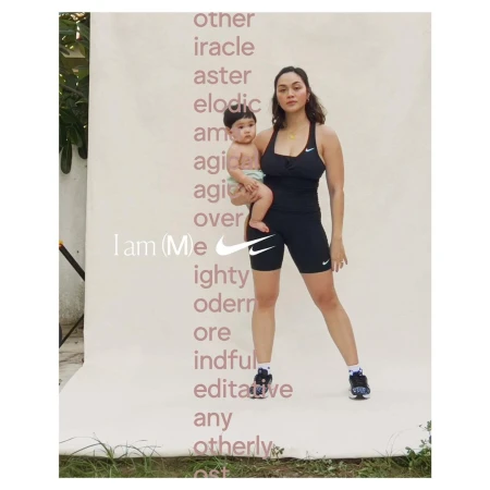 Mother holds her baby against a branded backdrop in a fashion-style shot. She's wearing black athletic wear and tennis shoes.