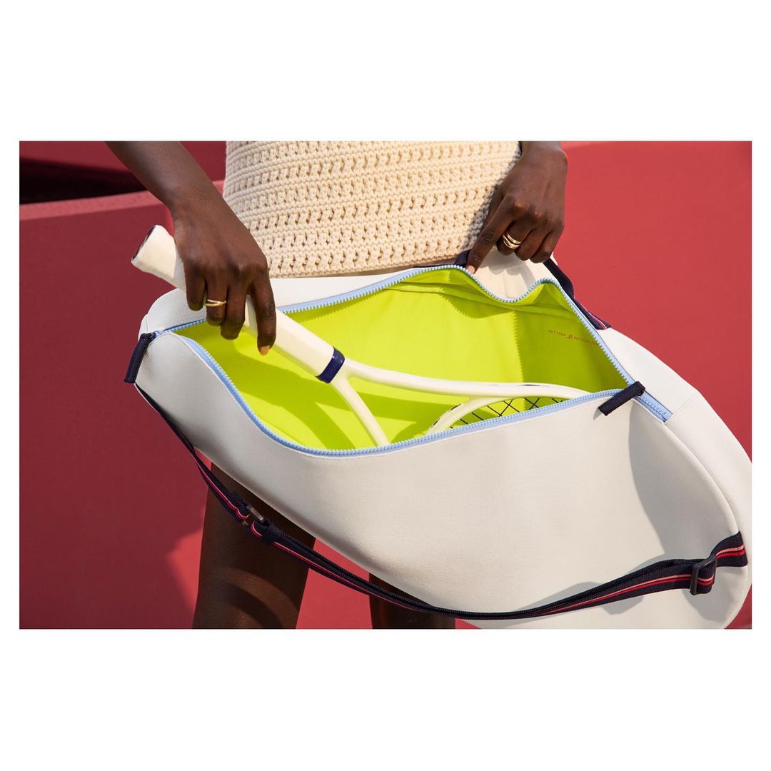 Woman opens a white tennis bag, revealing a neon yellow interior, containing a tennis racket, against a pink background.