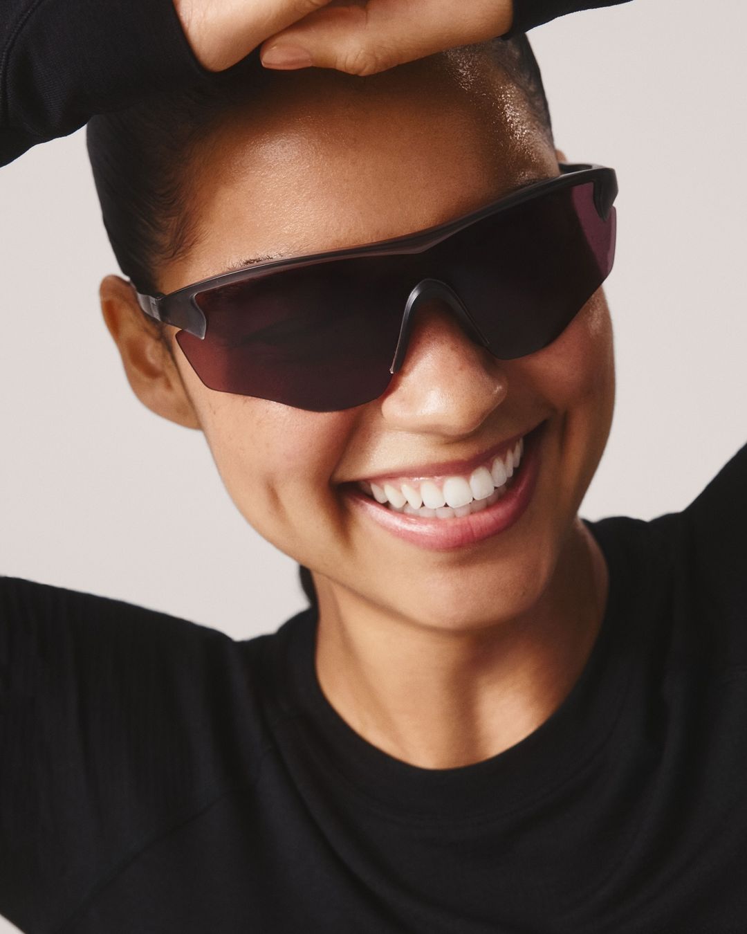 Close-up studio shot of a woman with dark hair smiling and wearing black sunglasses and a black top against a light background.