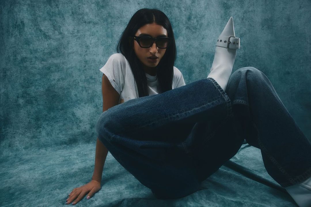 Woman wearing sunglasses, white t-shirt, and blue jeans, holding a white pointed-toe heeled boot up toward the camera against a textured teal backdrop