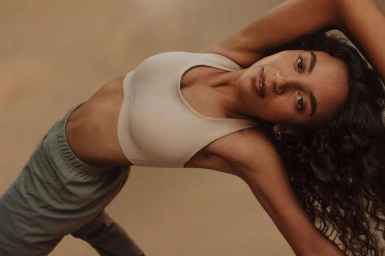 Woman in a beige sports bra and grey trousers stretching outdoors with arms raised