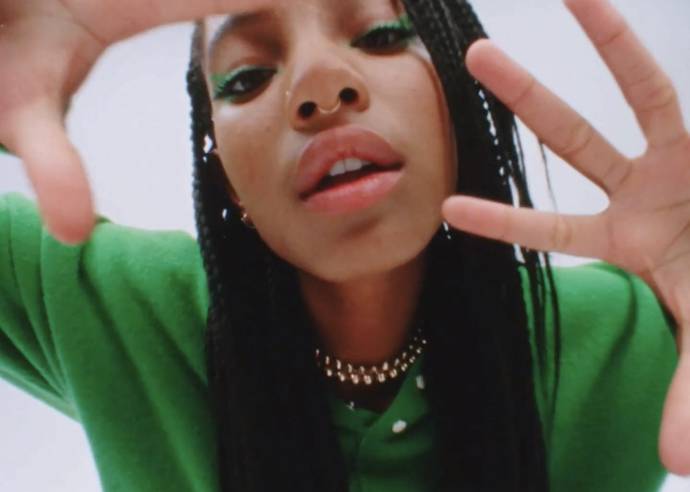 Eye-level close-up of a stylish adult female with green eyeshadow and braided hair in a green shirt, posing with hands framing the face.
