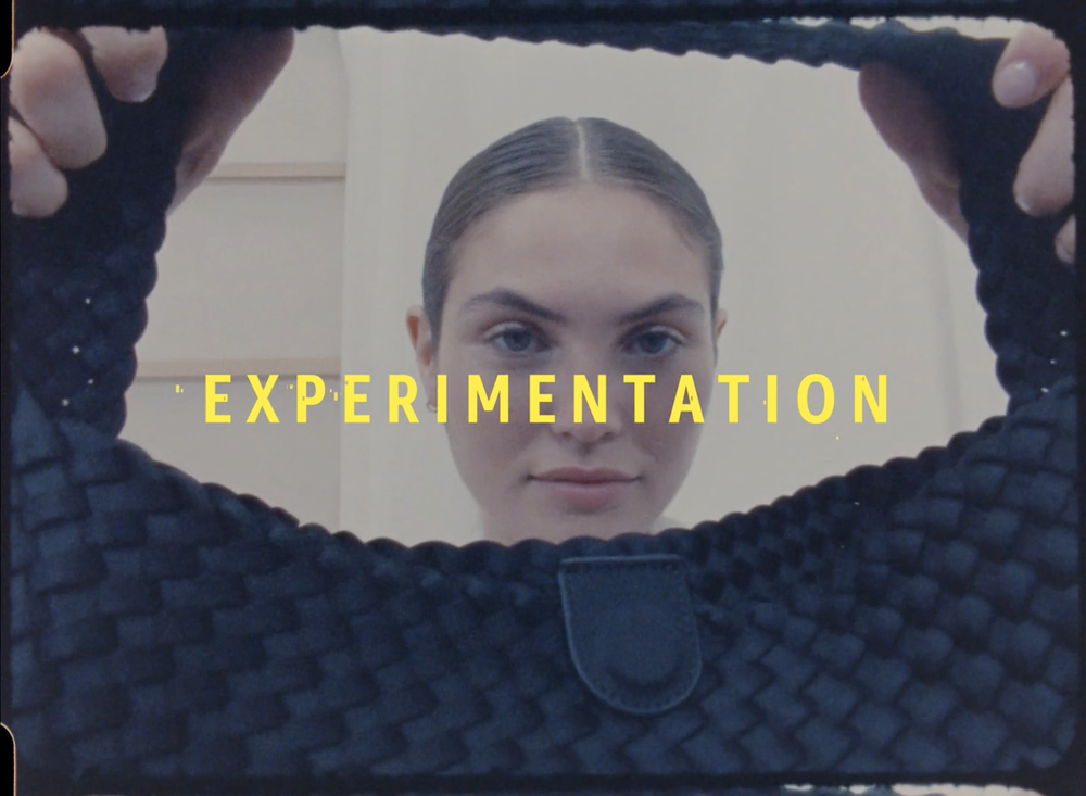 Woman's face framed by dark woven handbag with yellow text overlay 'EXPERIMENTATION' in a studio setting with neutral wall.