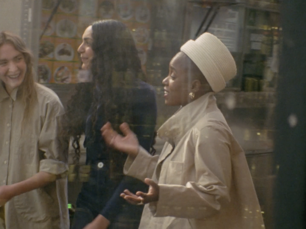 Three women standing indoors near shelves, one wearing a cream-colored trench coat and a beige hat, the other two in casual attire.