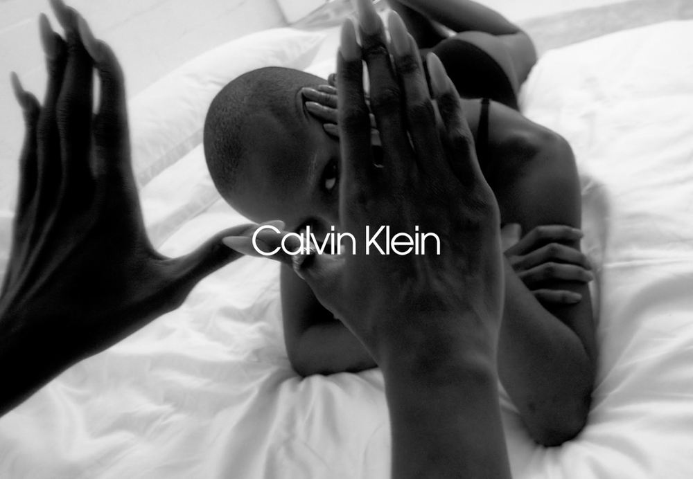 Striking black and white fashion image of a woman with a shaved head lying on a white bed, hands framing her face, Calvin Klein logo.
