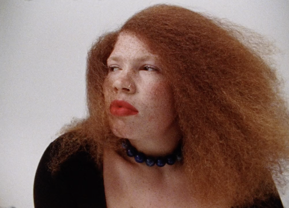 Portrait of a freckled red-haired woman with bold red lipstick and a blue beaded necklace, looking away against a neutral backdrop.