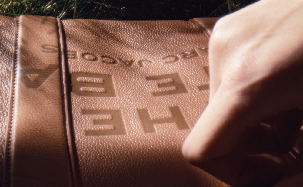 Close-up shot of a light brown leather bag with embossed 'MARC JACOBS' branding, an adult human hand, and green foliage visible in the background.