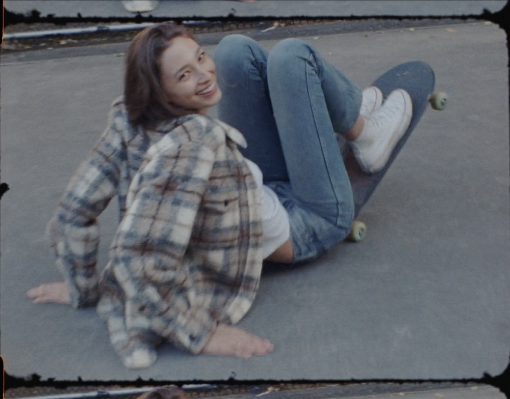 Smiling young woman poses barefoot on a skateboard wearing jeans and a plaid shirt with a concrete surface visible.