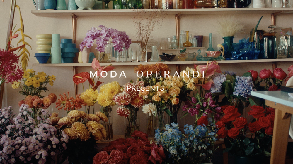 Abundant floral arrangement featuring roses, dahlias, and hydrangeas in an indoor setting, displayed on shelves alongside decorative glass vases.