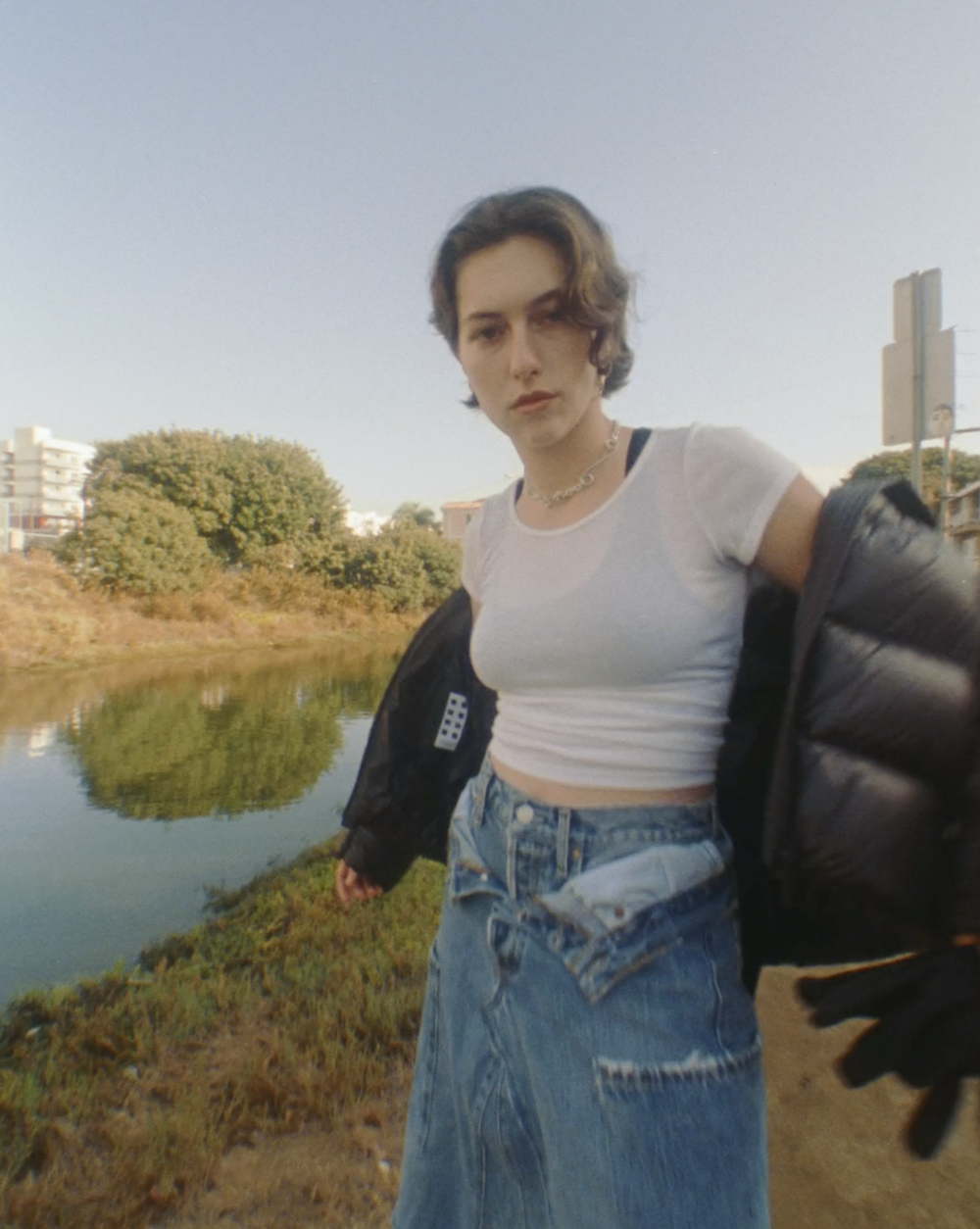 A stylish young woman in a white t-shirt and denim skirt stands by a river with trees on a sunny day, wearing a black jacket.