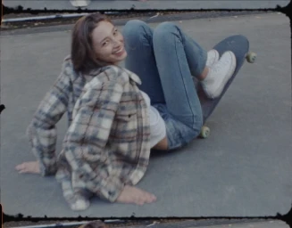 Smiling woman in a plaid jacket and blue jeans sitting on a skateboard outdoors with film borders visible.