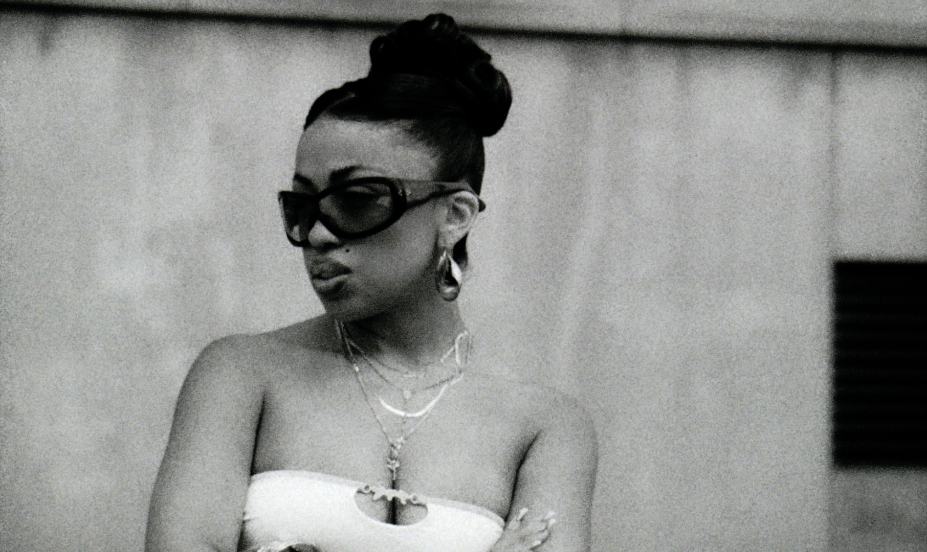 Monochrome portrait of a stylish adult woman wearing a strapless top, sunglasses, and layered jewelry against a textured wall.