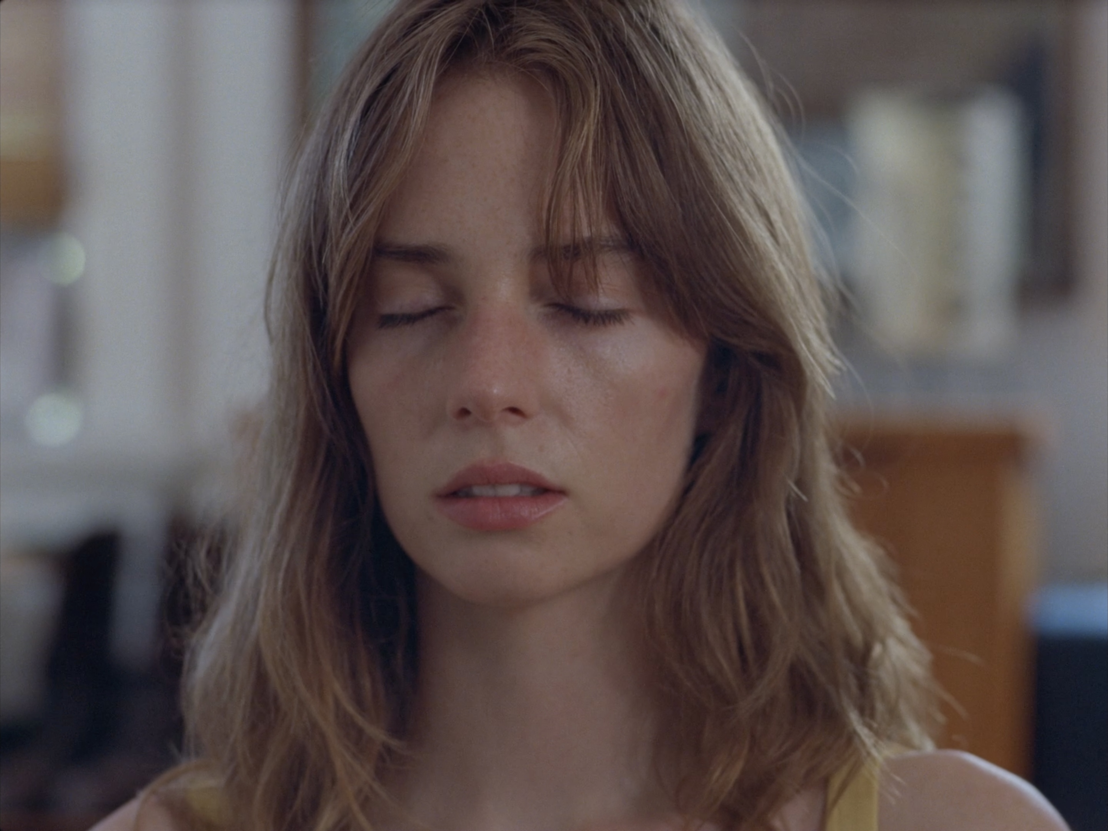 Close-up portrait of a young adult woman with light brown hair, eyes closed, fair skin, and a serene expression in an indoor setting.
