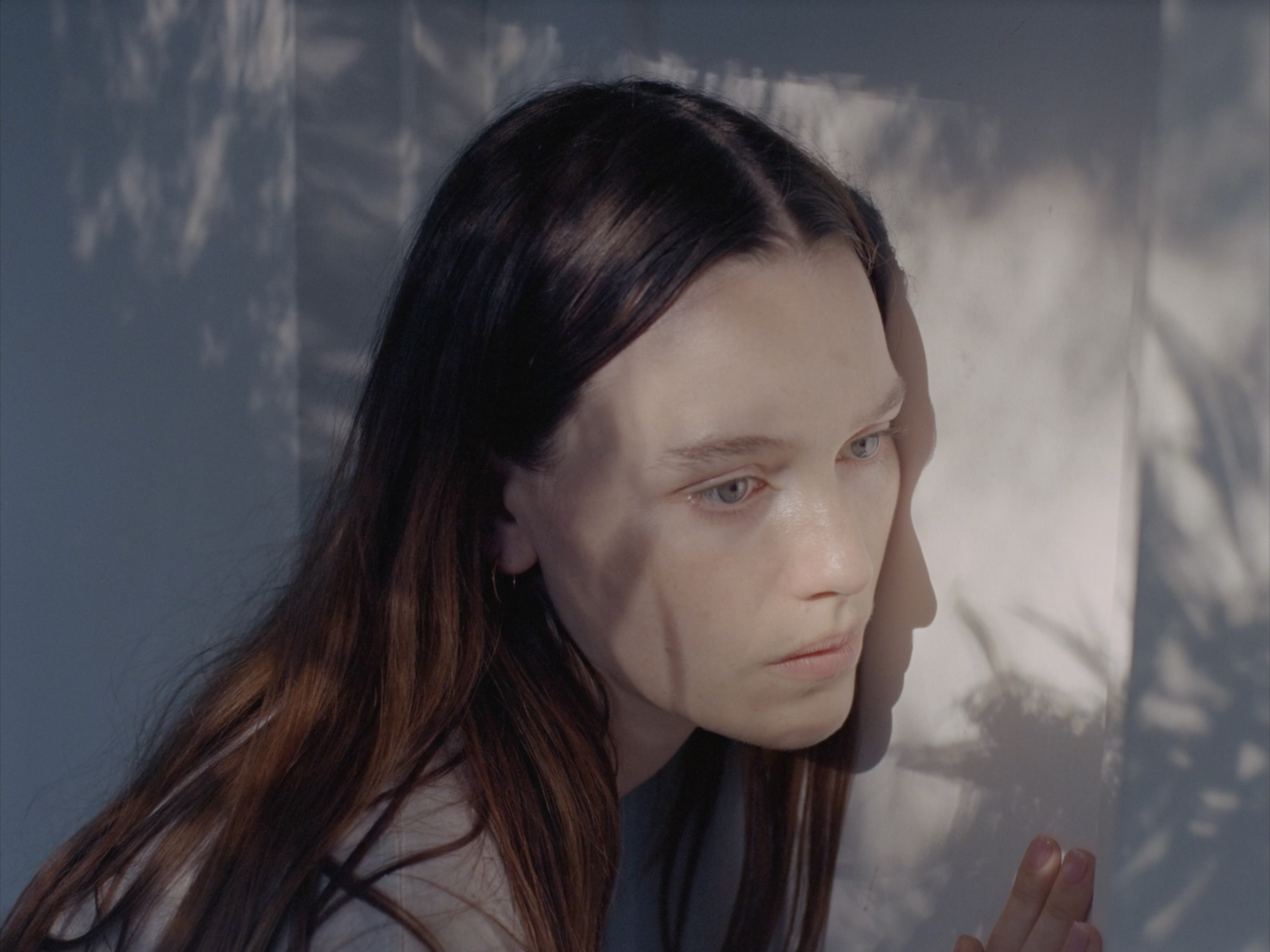 Portrait of a young adult woman with long brown hair looking away, standing indoors with sunlight and shadow patterns on the wall.