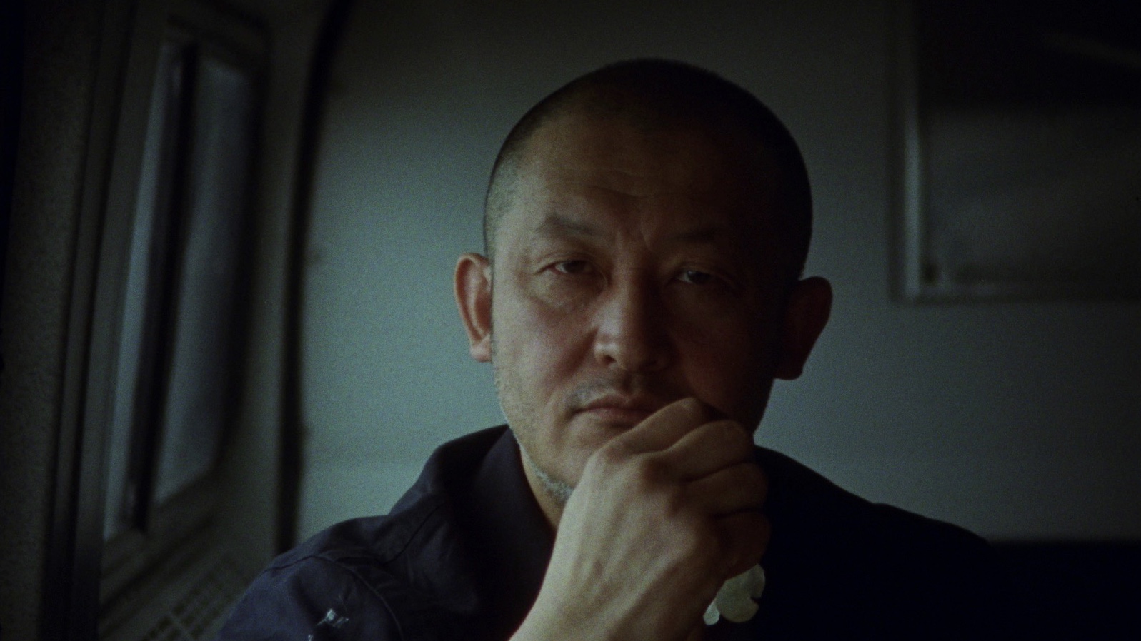 Close-up portrait of an Asian adult male with a shaved head, hand on chin, wearing a dark shirt, in dimly lit indoor setting.