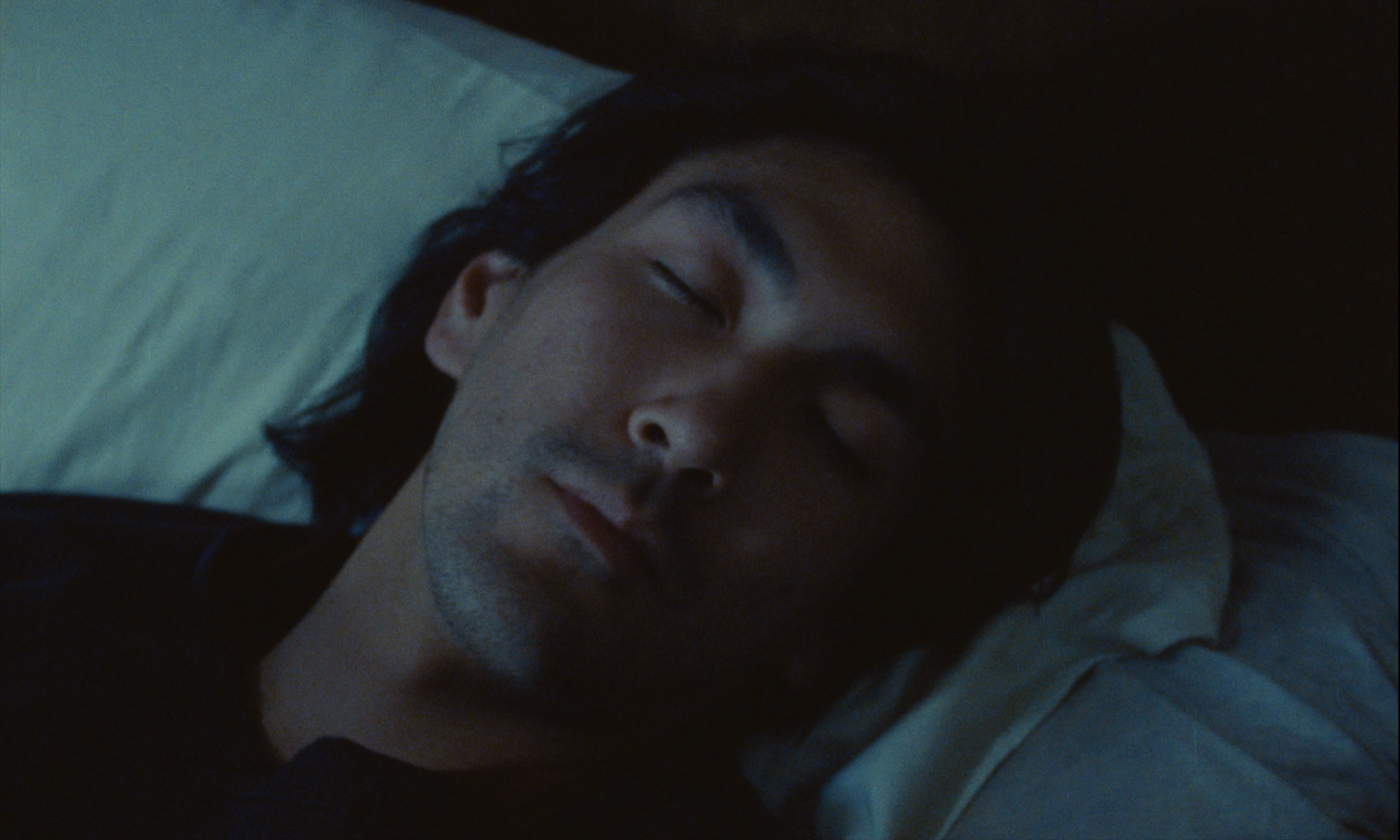Close-up of a sleeping adult male with long hair lying on a white pillow indoors in dim lighting.