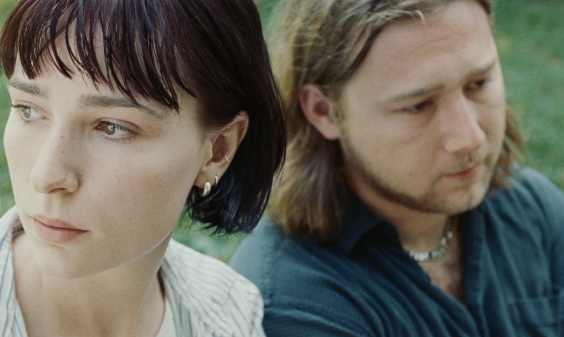 Close-up of a somber-looking woman with short dark hair and a man with long blond hair. They sit outside surrounded by a green grass background.