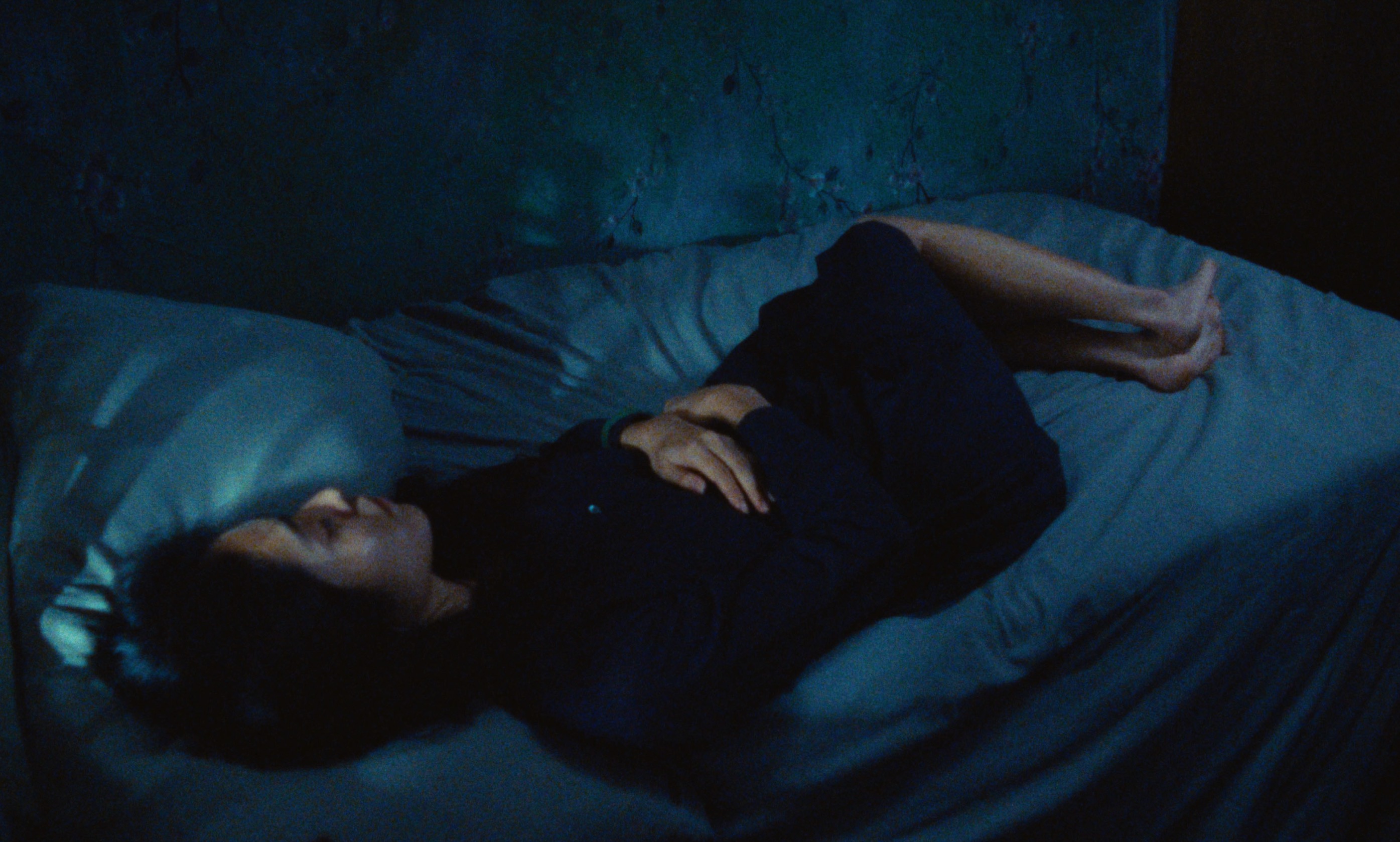 Adult female in dark dress lies on her side on a bed with a gray-blue textured wall in the background in a low-light, indoor bedroom.