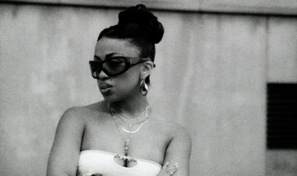 Black woman wearing sunglasses and layered necklaces with her hair in a high updo against a stone wall