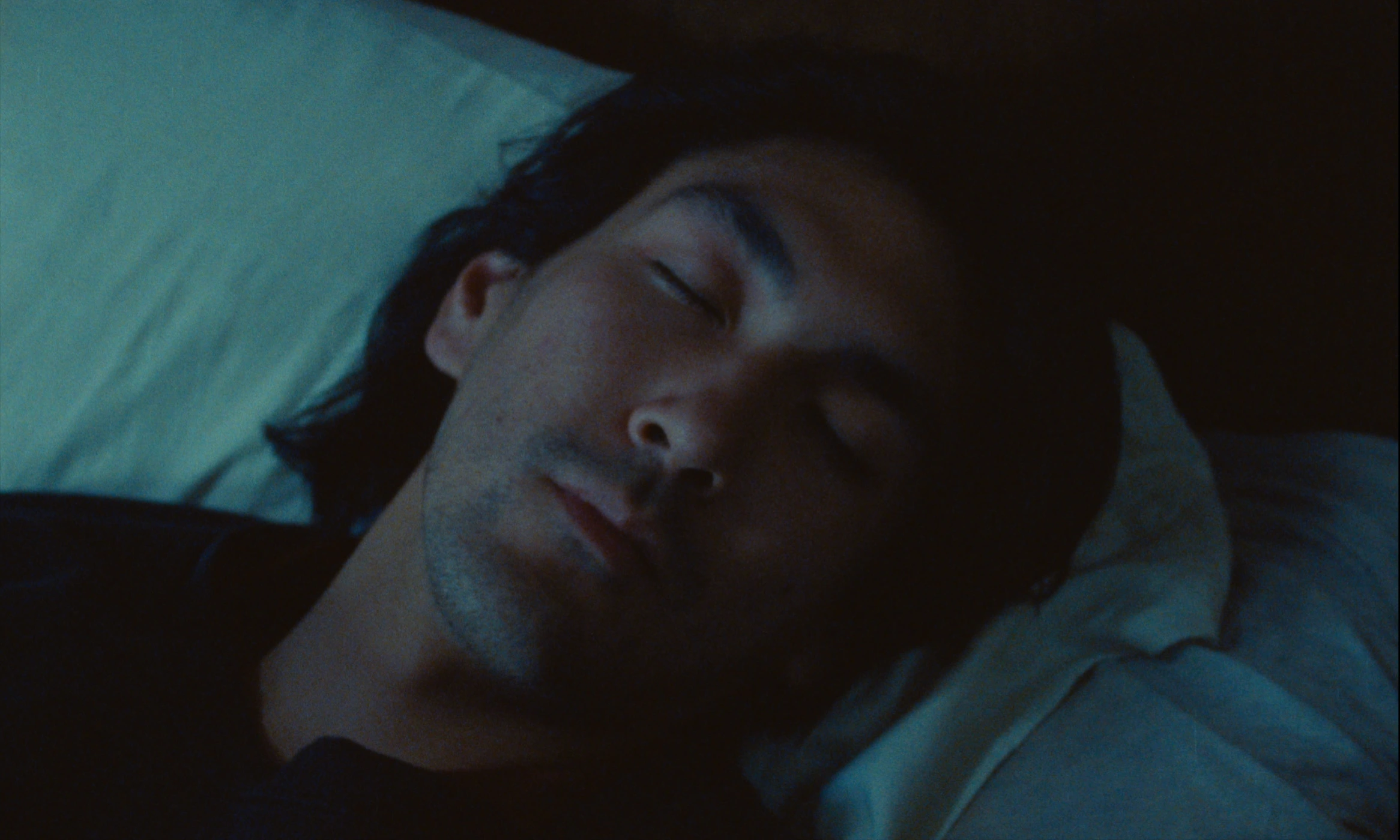 Close-up cinematic shot of a man sleeping with eyes closed, resting on white pillows in dim light.