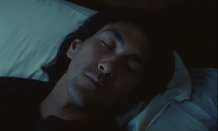 Close-up cinematic shot of a man sleeping with eyes closed, resting on white pillows in dim light.