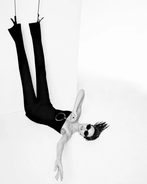 Upside down shot of a woman in a black bodysuit and sunglasses suspended by ropes in a white photo studio, black and white photography.