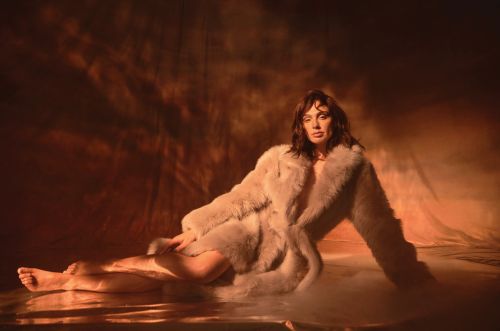 A woman in a fur coat reclines in a studio, bathed in warm light, creating a glamorous and intimate portrait. Soft, diffused lighting emphasizes texture.