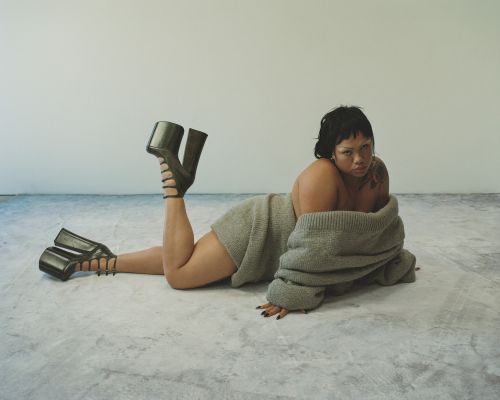 Woman lies on floor wearing a grey sweater and black, open-toed platform heels against a plain grey and white studio backdrop.