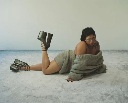 Woman lies on floor wearing a grey sweater and black, open-toed platform heels against a plain grey and white studio backdrop.