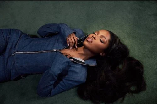 Woman in a denim jumpsuit reclines on a green surface, gazing upward. Long black hair fans out against the green backdrop.