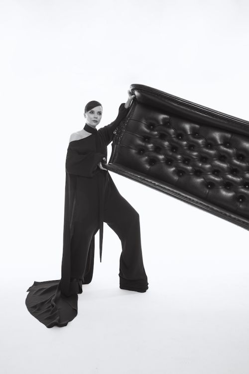 Woman in black dress poses with a tufted headboard in a studio setting, creating a fashion-forward and artistic portrait.