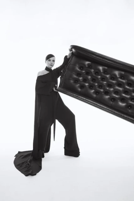 Woman in black dress poses with a tufted headboard in a studio setting, creating a fashion-forward and artistic portrait.